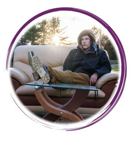 A person sits casually on a beige and brown leather couch placed incongruously in the middle of a quiet residential street. They wear a hooded winter jacket and brown trousers, with legs stretched out and feet resting on a glass coffee table positioned in front of the couch. The scene is bathed in the warm light of a setting sun, casting elongated shadows across the pavement. Surrounding the street are leafless trees, power lines, and suburban houses, all subtly illuminated by the golden hour glow. The juxtaposition of indoor furniture in an outdoor public space creates a surreal, contemplative atmosphere—blending domestic comfort with urban exposure. The image evokes themes of displacement, introspection, and the blending of private and public realms.