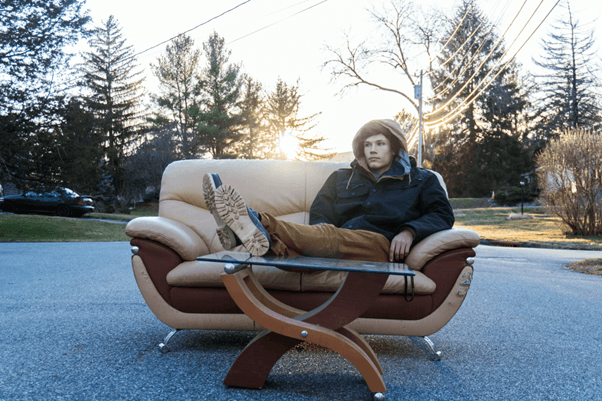 A person sits casually on a beige and brown leather couch placed incongruously in the middle of a quiet residential street. They wear a hooded winter jacket and brown trousers, with legs stretched out and feet resting on a glass coffee table positioned in front of the couch. The scene is bathed in the warm light of a setting sun, casting elongated shadows across the pavement. Surrounding the street are leafless trees, power lines, and suburban houses, all subtly illuminated by the golden hour glow. The juxtaposition of indoor furniture in an outdoor public space creates a surreal, contemplative atmosphere—blending domestic comfort with urban exposure. The image evokes themes of displacement, introspection, and the blending of private and public realms.