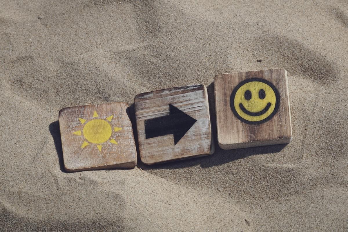 Three wooden tiles rest on sand: the left tile shows a yellow sun, the middle tile displays a black arrow pointing right, and the right tile features a yellow smiley face. The arrangement suggests a symbolic journey from brightness to positivity, set against a natural, earthy backdrop. Three wooden tiles rest on sand: the left tile shows a yellow sun, the middle tile displays a black arrow pointing right, and the right tile features a yellow smiley face. The arrangement suggests a symbolic journey from brightness to positivity, set against a natural, earthy backdrop.