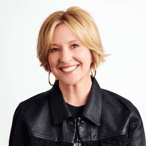 A professional portrait of Brené Brown, renowned author and researcher known for her work on vulnerability, courage, and shame. She is smiling warmly against a plain white background, wearing a black leather jacket and large hoop earrings. Her short blonde hair is styled neatly, and her expression conveys approachability and confidence. The clean backdrop and direct gaze suggest the image is intended for promotional or identification use, reflecting her public persona as a thought leader in emotional intelligence and personal growth. The composition is simple yet impactful, emphasisng authenticity and presence