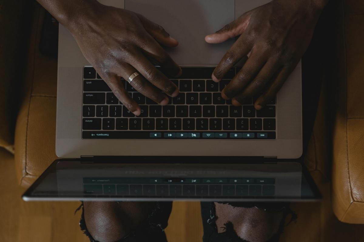 A top-down close-up view of a person actively using a laptop, likely a MacBook, identifiable by its keyboard layout and touch bar. The individual’s hands rest on the keyboard in a poised typing position, suggesting engagement with work, study, or communication. The screen reflects the keyboard and hands, adding depth and symmetry to the composition. The person wears a ring on their left hand and torn black jeans, introducing subtle personal style elements. The setting appears casual and modern, with the chair and posture indicating a focused yet relaxed digital interaction. The image captures a moment of contemporary tech use, emphasizing themes of productivity, individuality, and the tactile nature of digital engagement