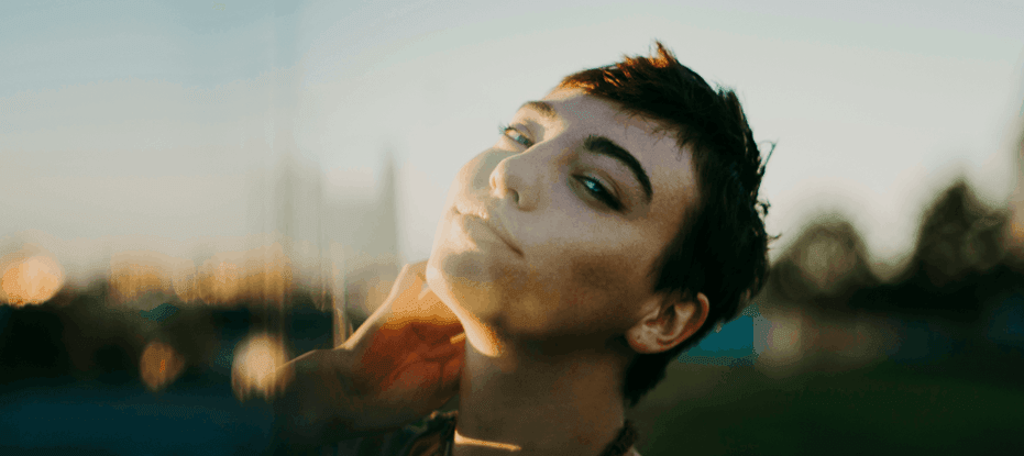 A close‑up portrait of a person outdoors during golden hour, their head tilted slightly back and to the side with one hand resting gently on their neck. Soft, warm sunlight illuminates their face, creating a gentle glow. The blurred background shows hints of greenery and sky, giving the image a serene, contemplative atmosphere