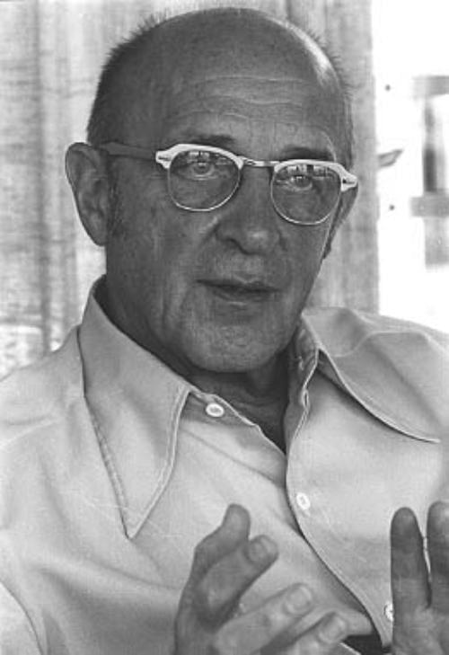 A black-and-white photograph of Carl Rogers, the influential humanistic psychologist, captured in a candid moment of expression. He wears glasses and a light-colored button-up shirt, seated indoors with curtains visible in the background. His hands are raised mid-gesture, suggesting he is engaged in conversation, explanation, or teaching. The grayscale tones emphasize the texture of his skin and the thoughtful intensity of his posture. The image conveys a sense of warmth, wisdom, and attentiveness—qualities central to Rogers’ person-centered approach. The composition highlights his role as a communicator and educator, making the image suitable for contexts related to psychology, counselling, or historical reflection on therapeutic practice