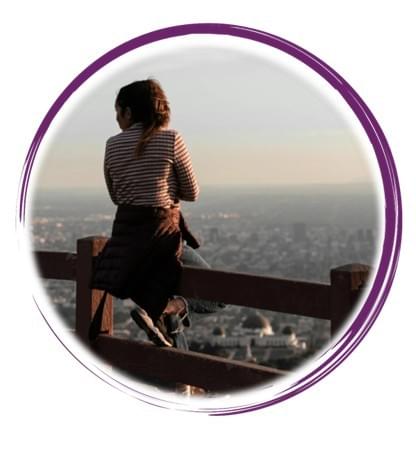 A casually dressed person sits alone on a wooden fence at a scenic overlook, gazing out over a sprawling cityscape below. The individual wears a striped shirt, jeans, and sneakers, with a dark jacket tied around their waist. A water bottle rests on the ground nearby, suggesting a pause during a walk or hike. The viewpoint is elevated—likely a hill or mountain—offering a panoramic view of densely packed urban buildings stretching toward the horizon. The sky above is hazy, casting a soft, muted light over the scene. The composition conveys a quiet moment of reflection and solitude, with the contrast between the natural foreground and the vast, structured city below evoking themes of contemplation, escape, and connection between nature and urban life