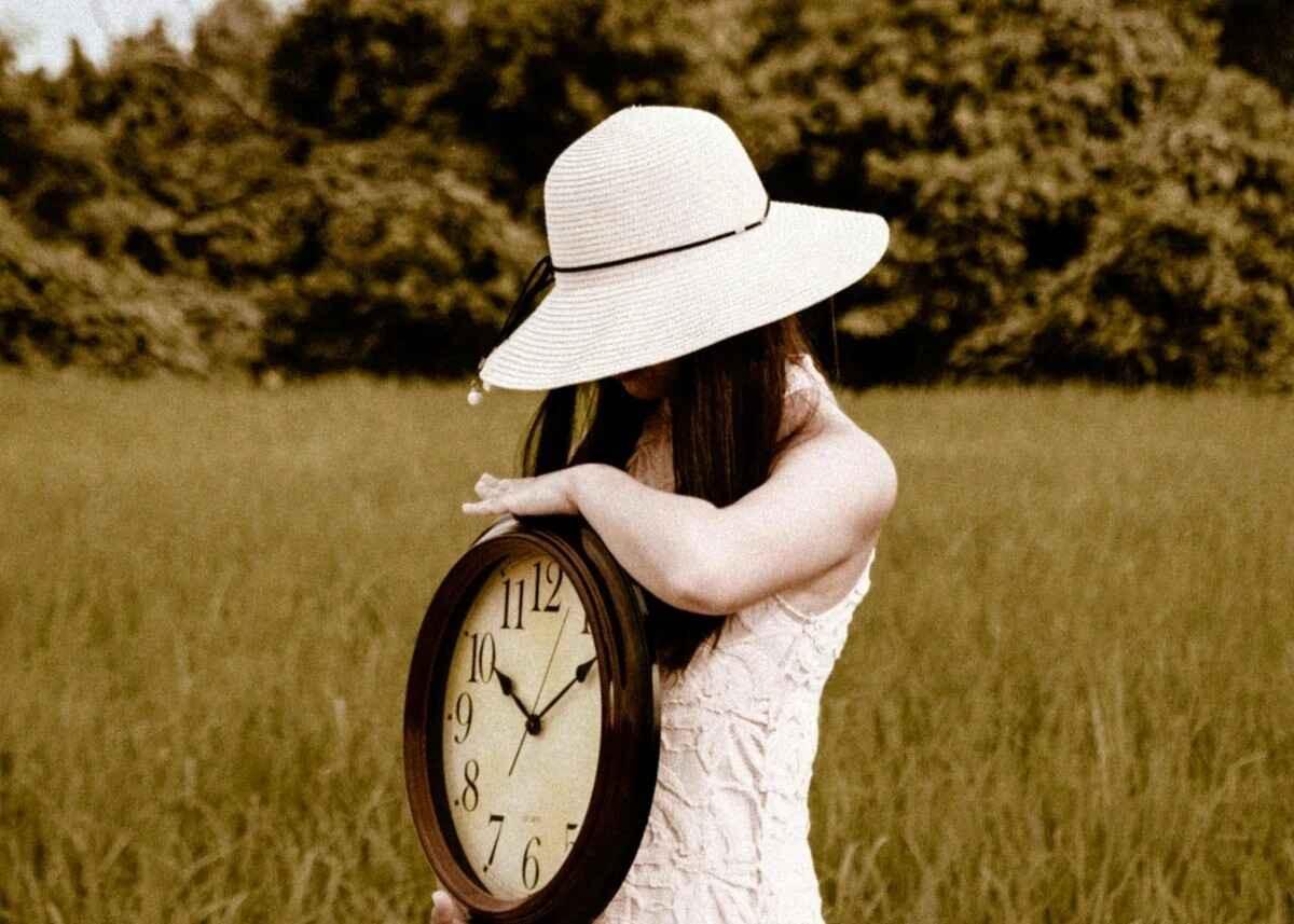 A person stands in a grassy field with dense trees behind them, wearing a light sleeveless dress and a wide‑brimmed hat that shades their face. They hold a large round clock close to their body with both arms resting on it, the hands pointing to 10:10. The image has a warm sepia tone, giving it a nostalgic, reflective atmosphere