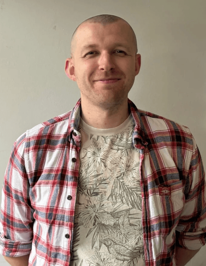 A person with short hair and a trimmed beard smiling warmly, wearing a light shirt and two necklaces, photographed against a plain light background. A person with short hair and a trimmed beard smiling warmly, wearing a light shirt and two necklaces, photographed against a plain light background.