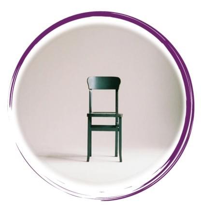 A single dark green wooden chair stands centered in a minimalist setting. The chair features a flat rectangular seat, four straight legs, and a slightly curved backrest composed of vertical slats. It is positioned on a smooth, light-colored floor that reflects soft shadows beneath and around the chair. The background is a neutral gradient, fading from light beige at the base to a soft gray toward the top, creating a calm and contemplative atmosphere. The lighting is gentle and directional, emphasizing the chair’s texture and form without harsh contrasts. The composition evokes themes of solitude, simplicity, and design focus, with the chair as the sole subject in an otherwise empty space.