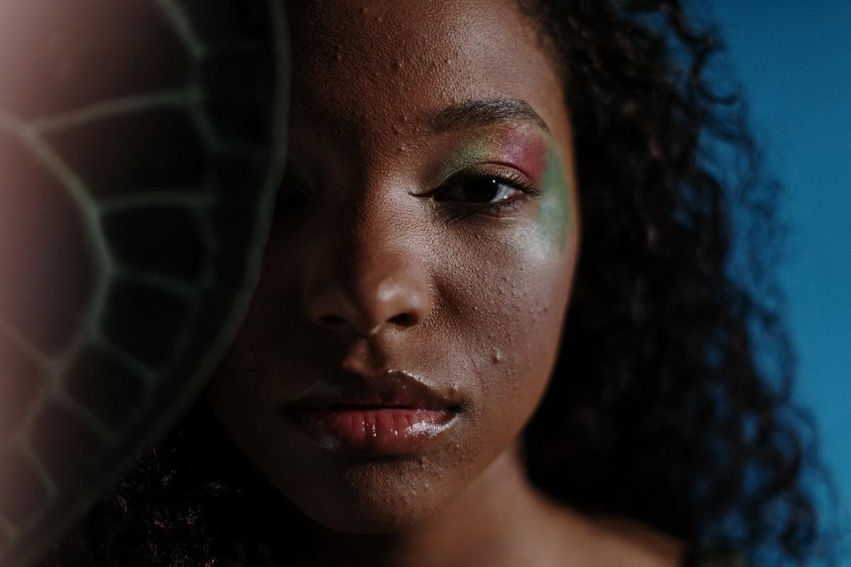 A close-up portrait of a person with curly hair set against a solid blue background. The individual’s face is partially obscured on the left side by a large leaf featuring a vivid green and white vein pattern, adding an organic, textural contrast. The person wears bold, colorful eye makeup—green and red tones blended across the eyelids—creating a striking visual focal point. Their skin is shown with natural texture, including visible pores and blemishes, emphasizing authenticity and realism. The composition balances vibrant color, natural imperfection, and botanical elements, evoking themes of identity, self-expression, and the interplay between human and nature