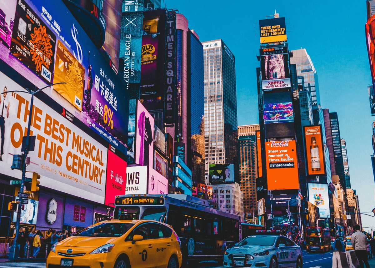 A busy Times Square street scene with tall buildings covered in bright electronic billboards, including ads for T‑Mobile, Coca‑Cola, Wicked, and other shows, with traffic such as a yellow taxi and a city bus in the foreground.