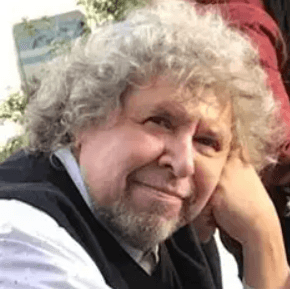 A candid outdoor portrait of Ernesto Spinelli, a distinguished figure in existential psychotherapy. He has curly gray hair and a beard, and is wearing a black vest over a white shirt. He rests his head gently on one hand and gazes slightly toward the camera with a thoughtful expression. The background includes trees and a sign, suggesting a natural or public setting. The image conveys quiet reflection and presence, with warm lighting and relaxed posture enhancing its contemplative tone.