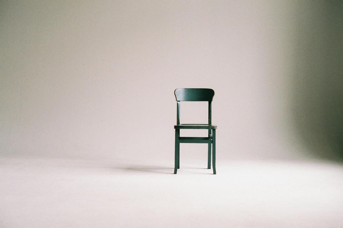 A single dark green wooden chair stands centered in a minimalist setting. The chair features a flat rectangular seat, four straight legs, and a slightly curved backrest composed of vertical slats. It is positioned on a smooth, light-colored floor that reflects soft shadows beneath and around the chair. The background is a neutral gradient, fading from light beige at the base to a soft gray toward the top, creating a calm and contemplative atmosphere. The lighting is gentle and directional, emphasizing the chair’s texture and form without harsh contrasts. The composition evokes themes of solitude, simplicity, and design focus, with the chair as the sole subject in an otherwise empty space.