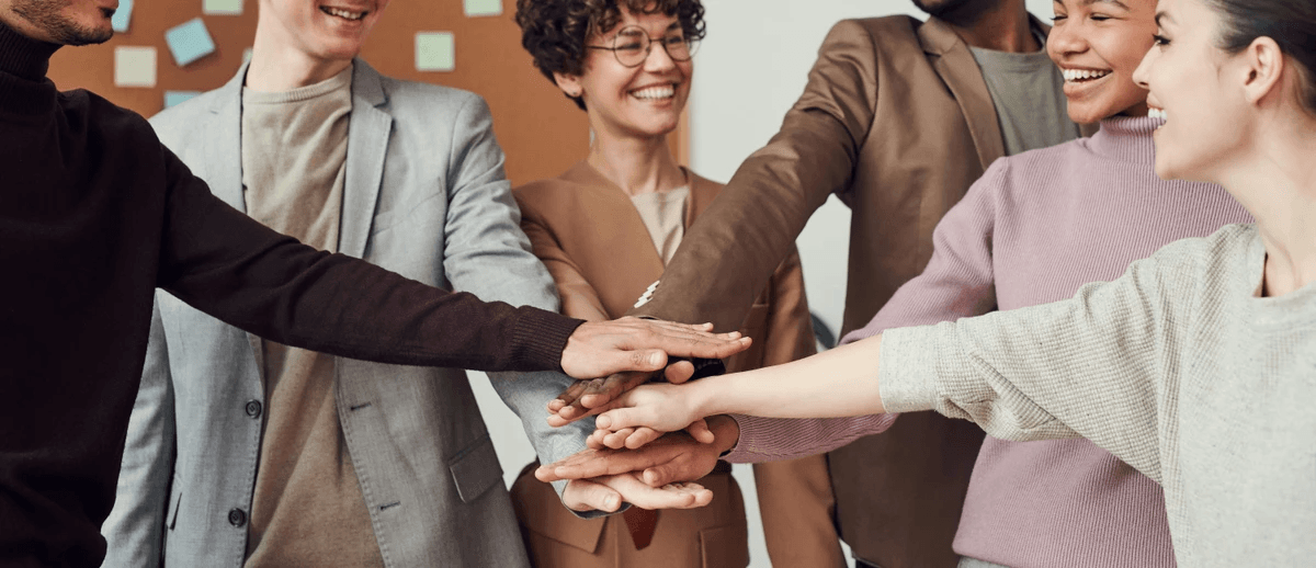 A group of people stand in a circle with their hands stacked together in the center, symbolizing unity, collaboration, and mutual support. The individuals are dressed in business casual attire, suggesting a professional or workplace setting. Their gesture conveys a shared commitment, possibly during a team-building exercise or a moment of collective affirmation. In the background, a corkboard with colorful sticky notes reinforces the office environment and hints at ongoing planning or brainstorming activities. The image captures the spirit of cooperation and positive interpersonal dynamics, emphasizing themes of teamwork, trust, and shared purpose in a professional context