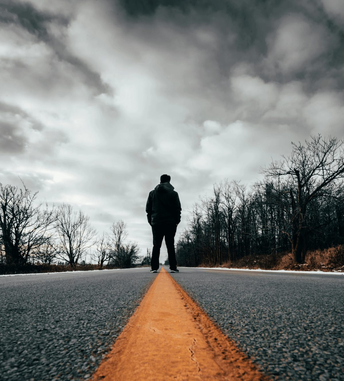A solitary figure stands on a deserted road, captured from a dramatic low-angle perspective that emphasizes the bold yellow centerline stretching toward the horizon. The person is dressed in dark clothing and faces away from the camera, gazing into the distance. Leafless trees line both sides of the road, suggesting a late autumn or winter setting. Above, the sky is filled with thick, overcast clouds that cast a moody, contemplative atmosphere across the scene. The composition uses strong leading lines and stark contrasts to evoke themes of solitude, introspection, and journey—both literal and emotional. The image’s visual tension between the grounded road and expansive sky adds depth and narrative potential