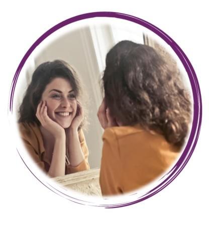 A person with wavy dark hair, dressed in a mustard-colored top, gazes into a large ornate mirror with a distressed white frame. They rest their face gently in both hands and smile warmly at their reflection, capturing a moment of candid joy and self-connection. The softly lit room behind them features white walls and a window that casts natural light across the scene, adding a sense of calm and intimacy. The composition emphasizes themes of self-acceptance, inner peace, and the quiet beauty of personal reflection.