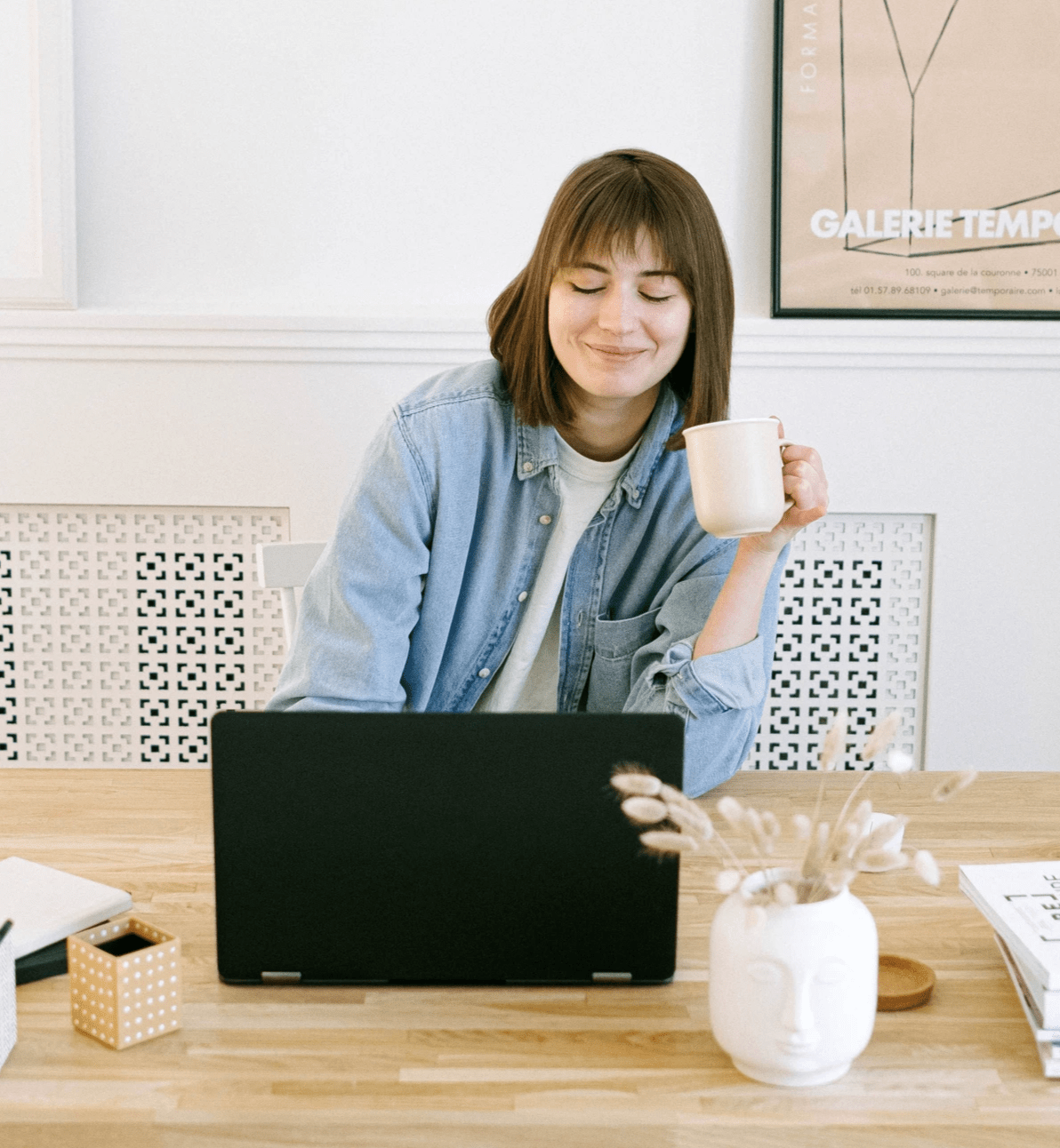 A person sits at a wooden table, working on a black laptop while holding a white mug in one hand. The setting is cozy and thoughtfully arranged: the tabletop includes a white ceramic vase filled with dried plants, a small container, and a closed notebook. Behind the person, a white wall features a framed poster with the words “GALERIE TEMP,” adding a touch of artistic flair. Soft natural light filters into the room, creating a calm and focused atmosphere. The scene evokes a sense of relaxed productivity, blending aesthetic comfort with quiet concentration—ideal for remote work, creative reflection, or therapeutic journaling.