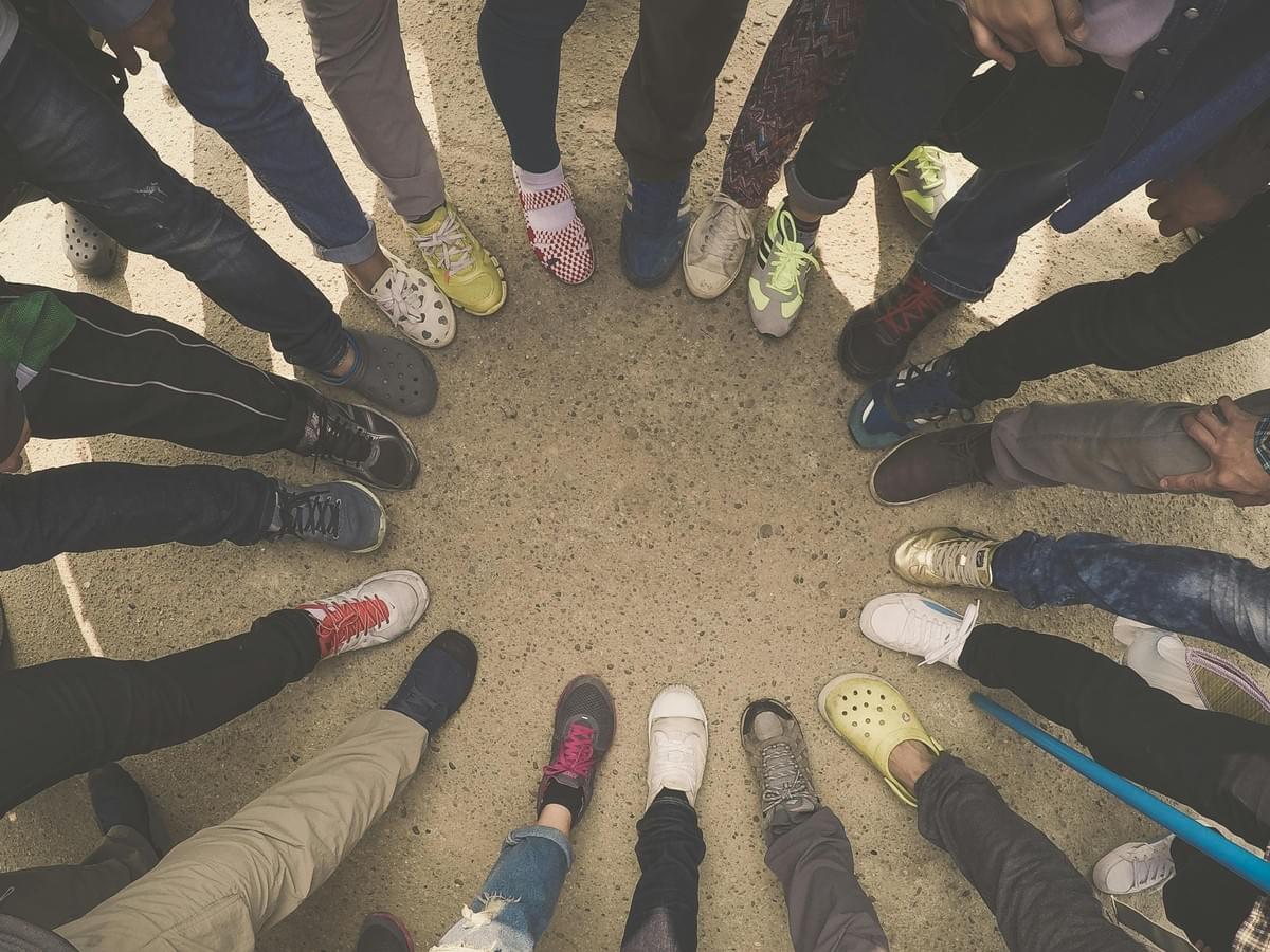 A group of people stand in a circle on a sandy or concrete surface, with only their legs and feet visible. Each person wears different footwear—sneakers, crocs, boots, and casual shoes—in varied colors and styles. Their feet point inward, forming a radial pattern that visually conveys diversity, unity, and individuality within a collective. A group of people stand in a circle on a sandy or concrete surface, with only their legs and feet visible. Each person wears different footwear—sneakers, crocs, boots, and casual shoes—in varied colors and styles. Their feet point inward, forming a radial pattern that visually conveys diversity, unity, and individuality within a collective.