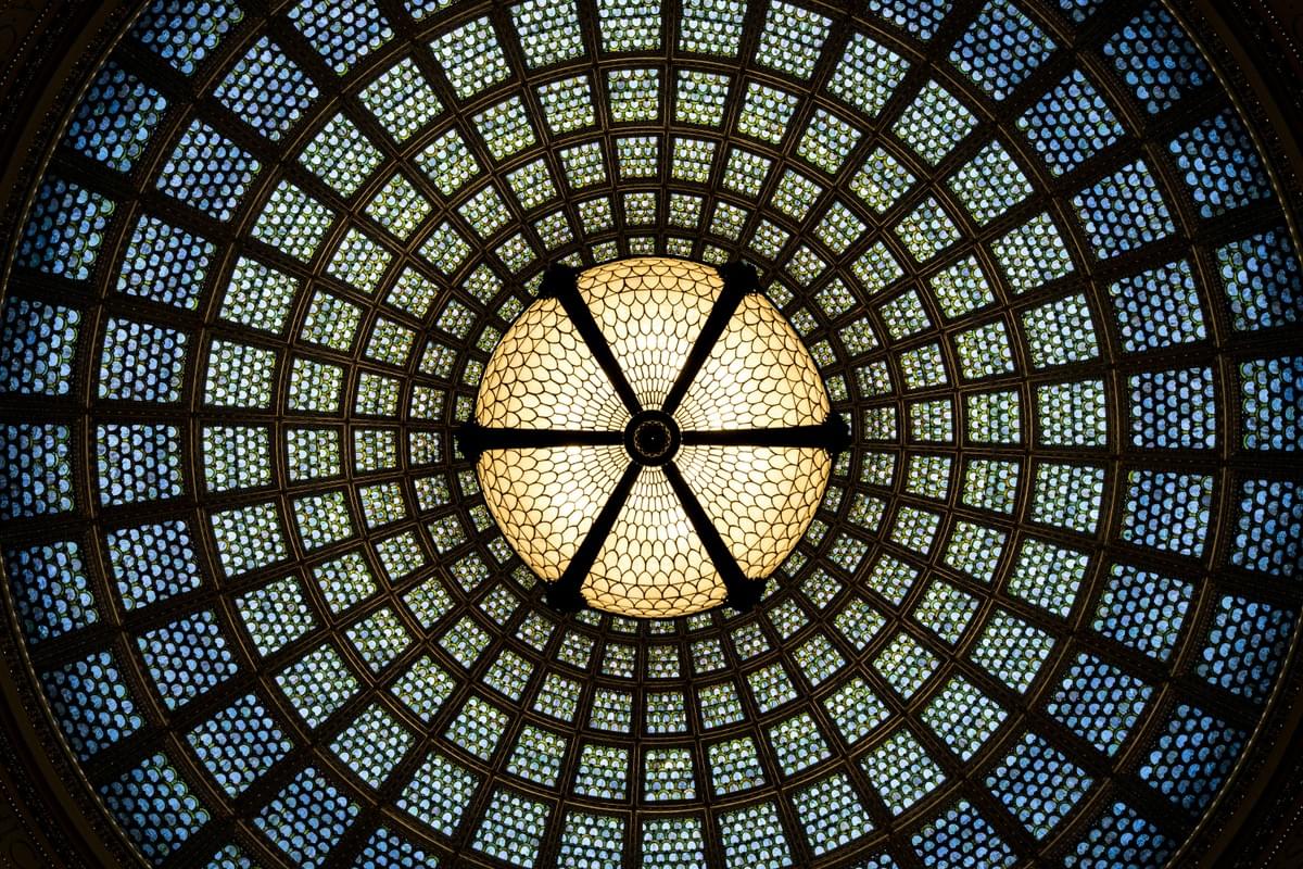 A symmetrical stained glass dome ceiling viewed from below, featuring concentric rings of hexagonal and circular glass panels. The center glows with warm yellow light, while the outer sections shift to cooler blue and green hues. A radial framework divides the segments, creating an intricate geometric pattern that highlights the dome’s craftsmanship and luminous beauty A symmetrical stained glass dome ceiling viewed from below, featuring concentric rings of hexagonal and circular glass panels. The center glows with warm yellow light, while the outer sections shift to cooler blue and green hues. A radial framework divides the segments, creating an intricate geometric pattern that highlights the dome’s craftsmanship and luminous beauty