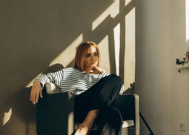 A person sits in a chair in a sunlit room, wearing a white shirt with black horizontal stripes and dark trousers. Their posture is relaxed, with one arm resting on the chair’s armrest and the other hand near their face. Sunlight streams through a nearby window, casting clear, angled shadows on the wall behind them. A small plant sits on a shelf to the right, adding to the calm, minimalistic atmosphere.