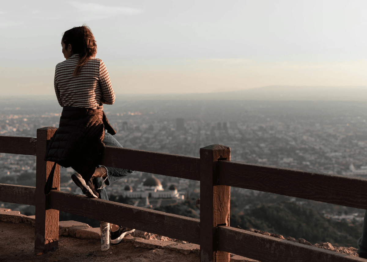 A casually dressed person sits alone on a wooden fence at a scenic overlook, gazing out over a sprawling cityscape below. The individual wears a striped shirt, jeans, and sneakers, with a dark jacket tied around their waist. A water bottle rests on the ground nearby, suggesting a pause during a walk or hike. The viewpoint is elevated—likely a hill or mountain—offering a panoramic view of densely packed urban buildings stretching toward the horizon. The sky above is hazy, casting a soft, muted light over the scene. The composition conveys a quiet moment of reflection and solitude, with the contrast between the natural foreground and the vast, structured city below evoking themes of contemplation, escape, and connection between nature and urban life