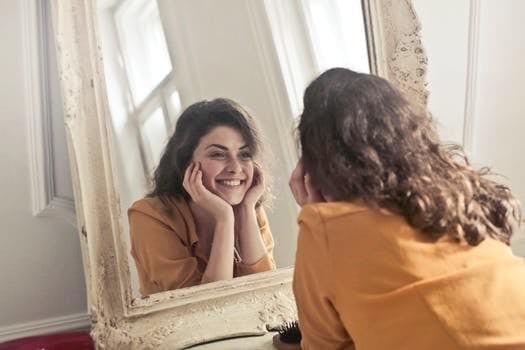 A person with wavy dark hair, dressed in a mustard-colored top, gazes into a large ornate mirror with a distressed white frame. They rest their face gently in both hands and smile warmly at their reflection, capturing a moment of candid joy and self-connection. The softly lit room behind them features white walls and a window that casts natural light across the scene, adding a sense of calm and intimacy. The composition emphasizes themes of self-acceptance, inner peace, and the quiet beauty of personal reflection.