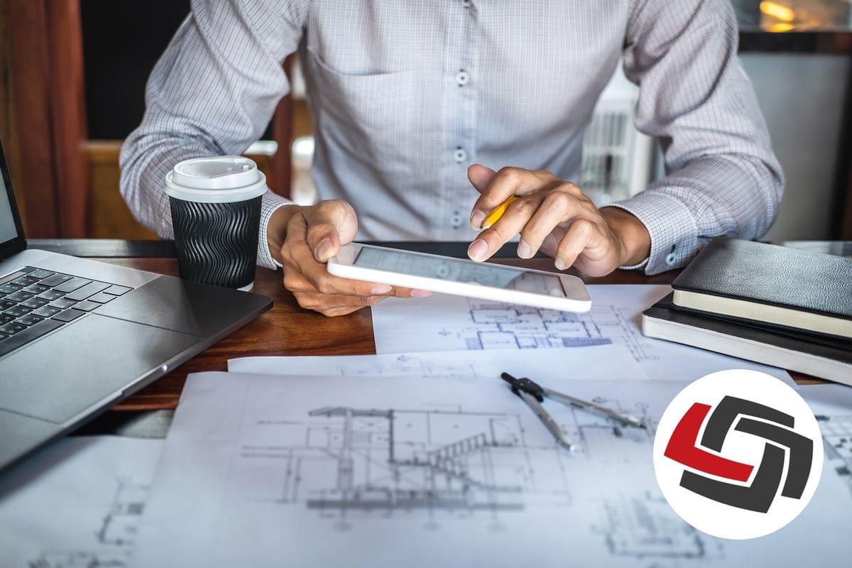 An architect is holding his mobile phone over his desk with architectural drawings, a coffee cup and a laptop on the desk
