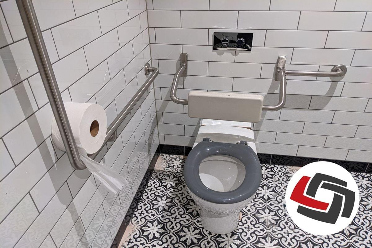 An accessible toilet in a corner of a room with a grey seat, grabrails and an ornate pattern on the tiled floor