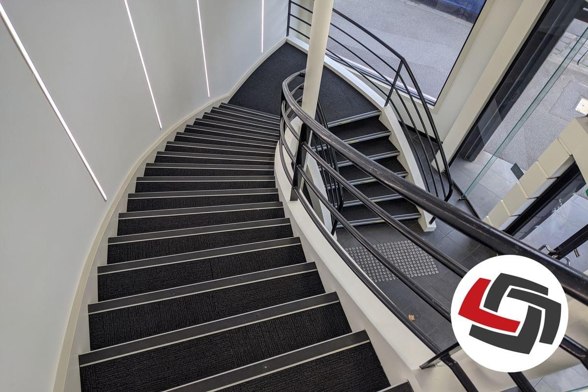 A view looking down a curved stairway with one black handrail, black carpet and black tread nosing strips and silver tactile indicators