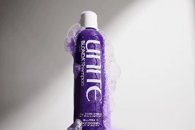 Purple Unite Blonda toning shampoo bottle covered in lavender foam and bubbles against light gray background, highlighting glossy texture. Purple Unite Blonda toning shampoo bottle covered in lavender foam and bubbles against light gray background, highlighting glossy texture.