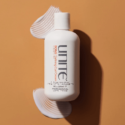 White Unite Boing curl defining cream bottle on warm tan surface with textured cream swipes above and below, showing smooth product finish. White Unite Boing curl defining cream bottle on warm tan surface with textured cream swipes above and below, showing smooth product finish.