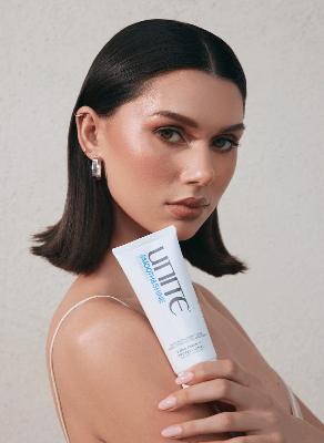 Person with sleek dark bob hair holds Unite Smooth & Shine cream tube against a neutral studio backdrop. Person with sleek dark bob hair holds Unite Smooth & Shine cream tube against a neutral studio backdrop.