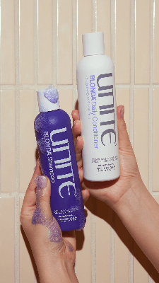 Person in white towel holding purple Unite Blonde Life brightening veil bottle near bathroom sink, preparing to dispense product. Person in white towel holding purple Unite Blonde Life brightening veil bottle near bathroom sink, preparing to dispense product.