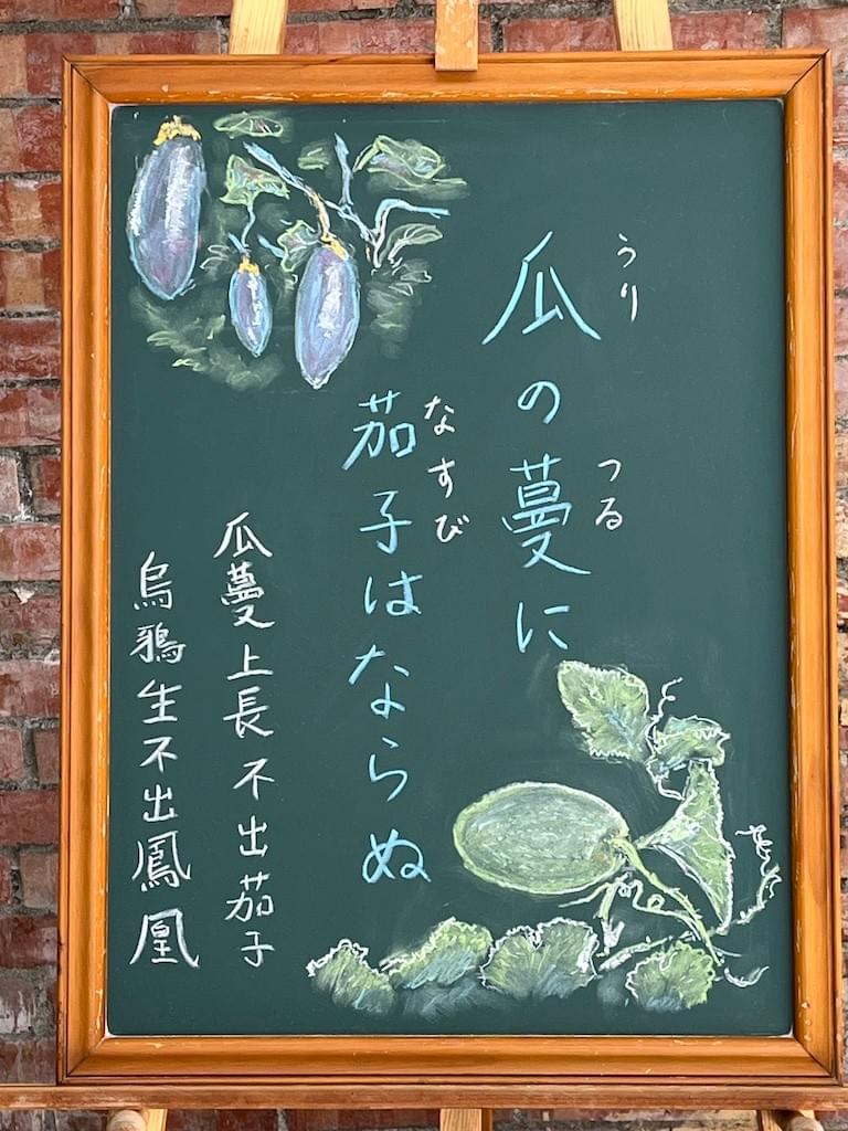 小さい黒板にチョークで絵が描いてあります。日本語のことわざ・慣用表現を紹介するための絵です。上部に棚の上の蔓からぶら下がっているナスのイラストが描いてあります。下部に蔓になっている瓜(マクワウリ)のイラストが描いてあります。中央に縦書き二行で「瓜の蔓に茄子はならぬ」と書いてあります。