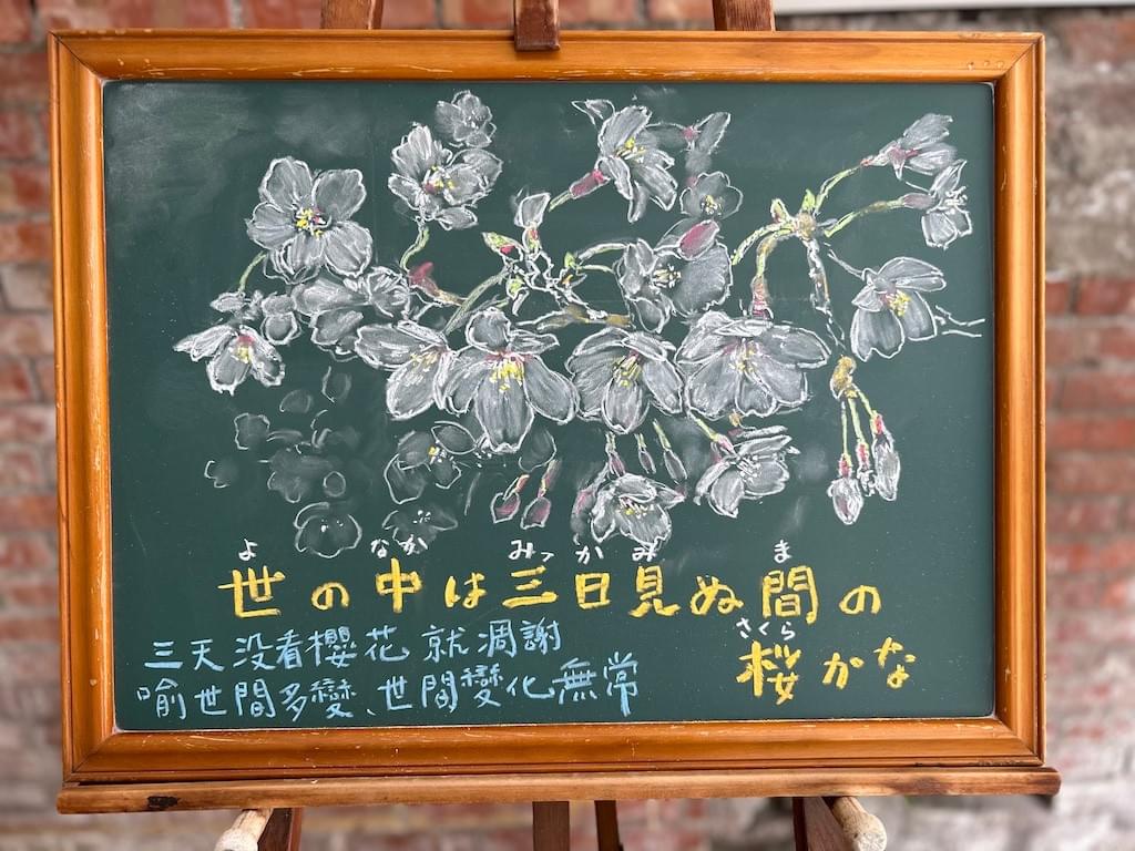 小さい黒板にチョークで絵が描いてあります。日本語のことわざ・慣用表現を紹介するための絵です。下部に横書きで「世の中は三日見ぬ間の桜かな」と書いてあり、上部に咲き誇る桜のイラストが描いてあります。