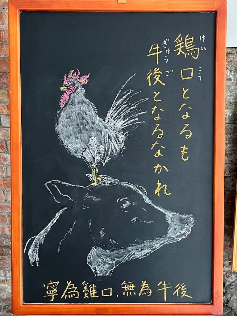 小さい黒板にチョークで絵が描いてあります。日本語のことわざ・慣用表現を紹介するための絵です。右上部に縦書きで、「鶏口となるも牛後となるなかれ」と書いてあり、下部に大きな牛の頭の絵、牛の目は上を向いています。そして牛の頭の上に白い鶏が立っているイラストが描いてあります。