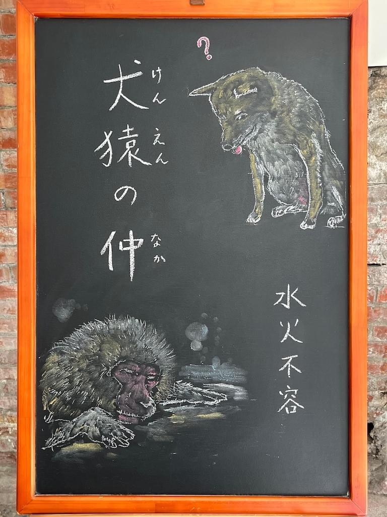 小さい黒板にチョークで絵が描いてあります。日本のことわざ・慣用表現を紹介するための絵です。左上に縦書きで「犬猿の仲」と書いてあり、その右に下を向いた柴犬のイラストが描いてあります。柴犬の視線の先、左下には温泉に入っている猿のイラストが描いてあります。猿は目をつぶっていて、気持ちよさそうです。