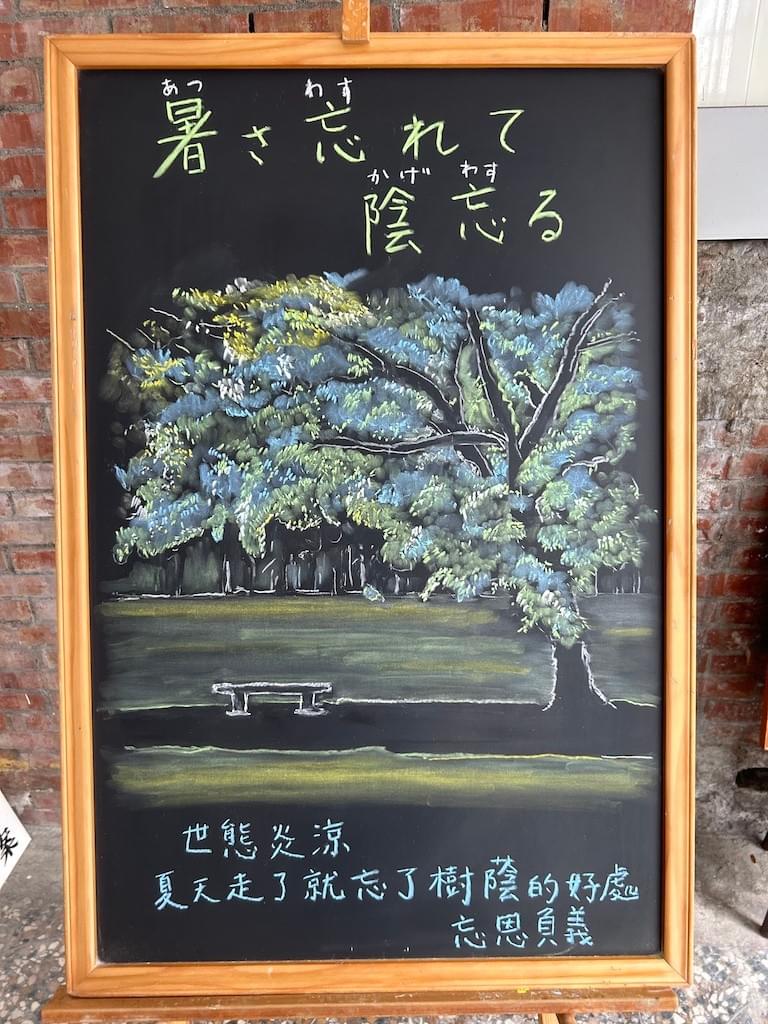 小さい黒板にチョークで絵が描いてあります。日本のことわざを紹介するための絵です。公園らしきところで、広い芝生が広がっているところ、右手前に大きな木があり、その下に木陰ができています。そこに一脚ベンチがあります。人は見えません。