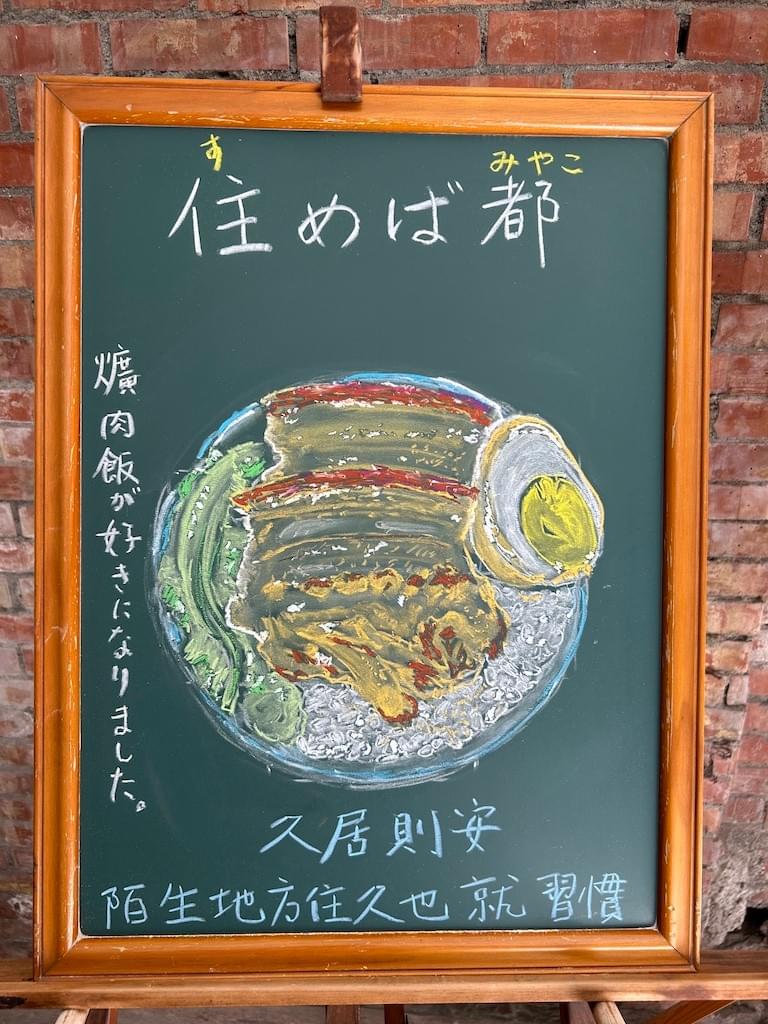 小さい黒板にチョークで絵が描いてあります。日本語のことわざ・慣用表現を紹介するための絵です。上部に横書きで「住めば都」とあり、下部に台湾の「爌肉飯」のイラストがあります。丸い器に白いご飯、その上にゆで卵、豚の角煮、茹でた青野菜が載っています。