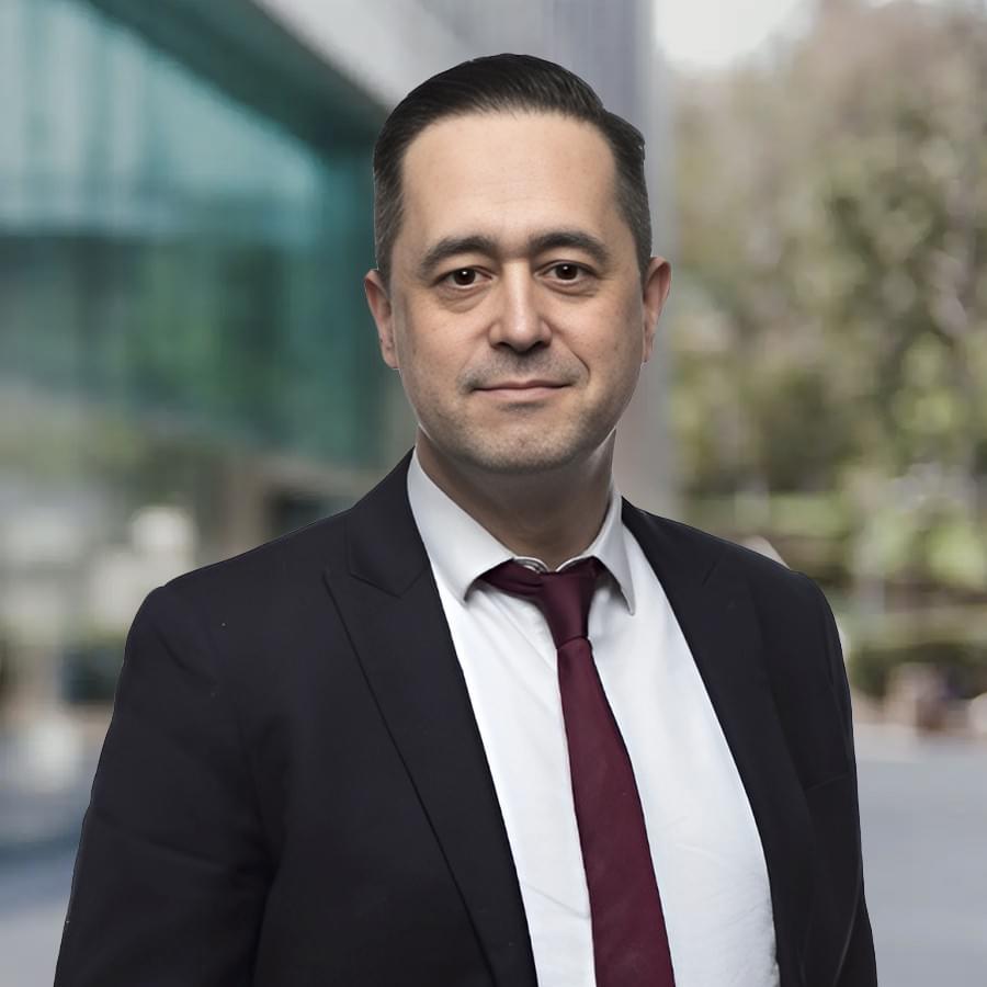 Mark Buzan in a dark suit and burgundy tie standing outdoors in front of a modern glass building, looking at the camera with a neutral expression.