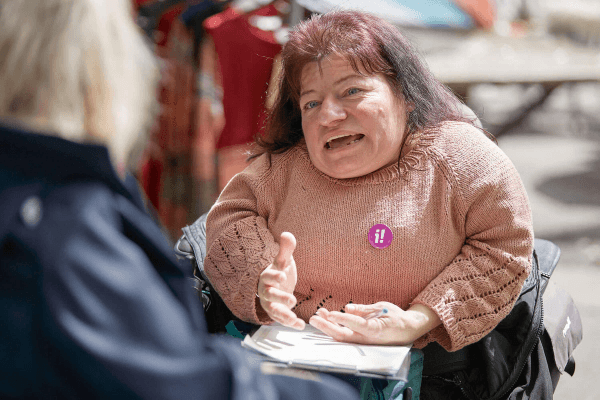 Eine Person mit Behinderung und Button der Inklusions-Initiative sammelt Unterschriften und erklärt der gegenüber sitzenden Person die Argumente für die Initiative.