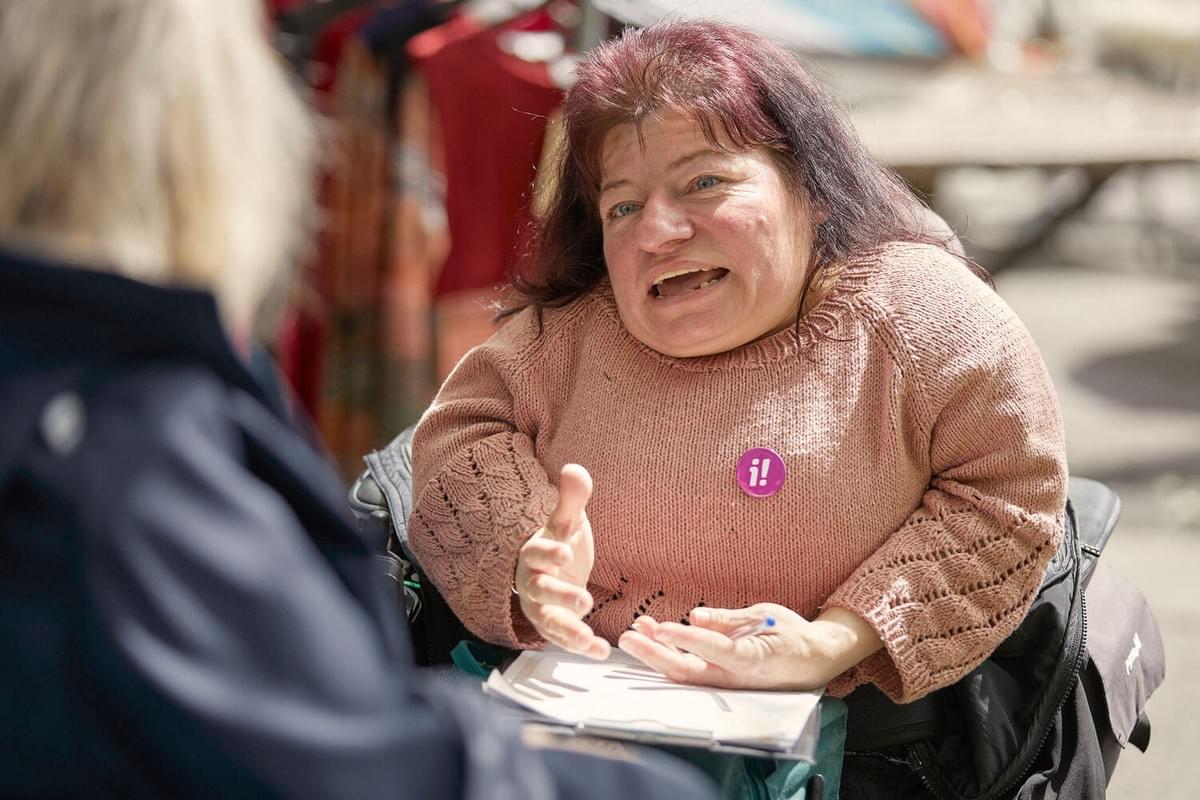 Eine Person mit Behinderung und Button der Inklusions-Initiative sammelt Unterschriften und erklärt der gegenüber sitzenden Person die Argumente für die Initiative.