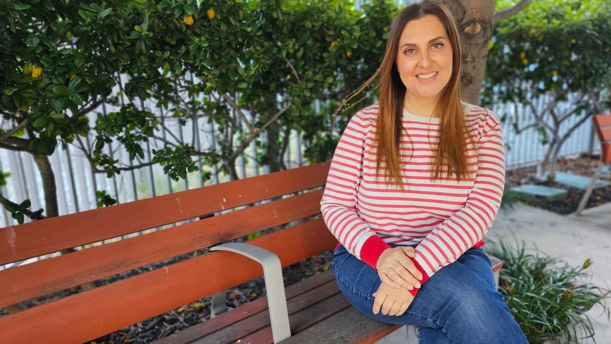 Kristyn Medeiros, host of the Flying Upstream podcast, sitting on a wooden bench outdoors surrounded by greenery, wearing a red and white striped long sleeve shirt and jeans, smiling calmly and confidently toward the camera.