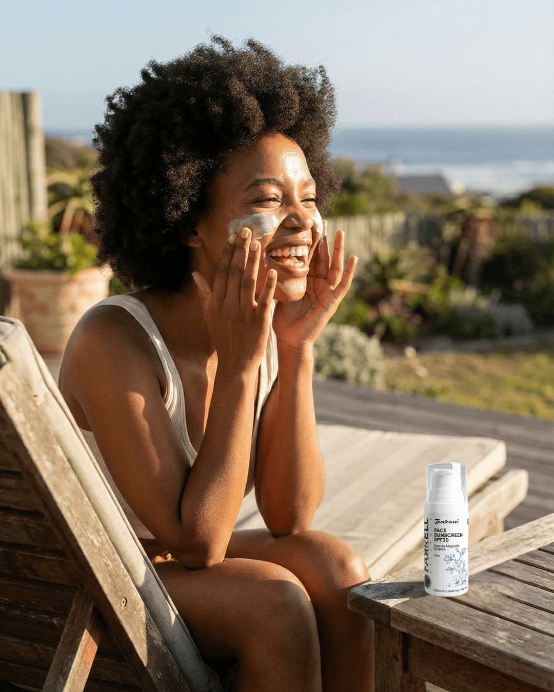 A smiling woman applies protective cream to her face while relaxing outdoors on a sunny deck with an ocean view. On the wooden table beside her sits a bottle of Naturallye Face Sunscreen SPF30, highlighting the product available on Glowlink.