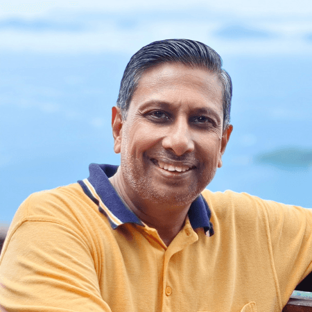 Portrait of a man with short black and grey hair and a trimmed beard, wearing a yellow T shirt and smiling while looking directly at the camera against a sky blue background."