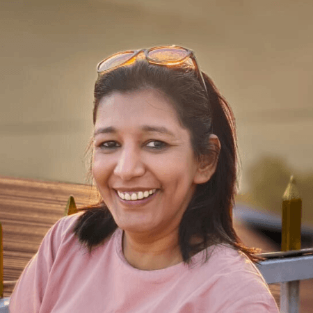 "Portrait of a woman with medium-length dark hair, wearing a pink top, smiling brightly while looking sideways at the camera. She is seated on a wooden bench with a warm sunset in the background.