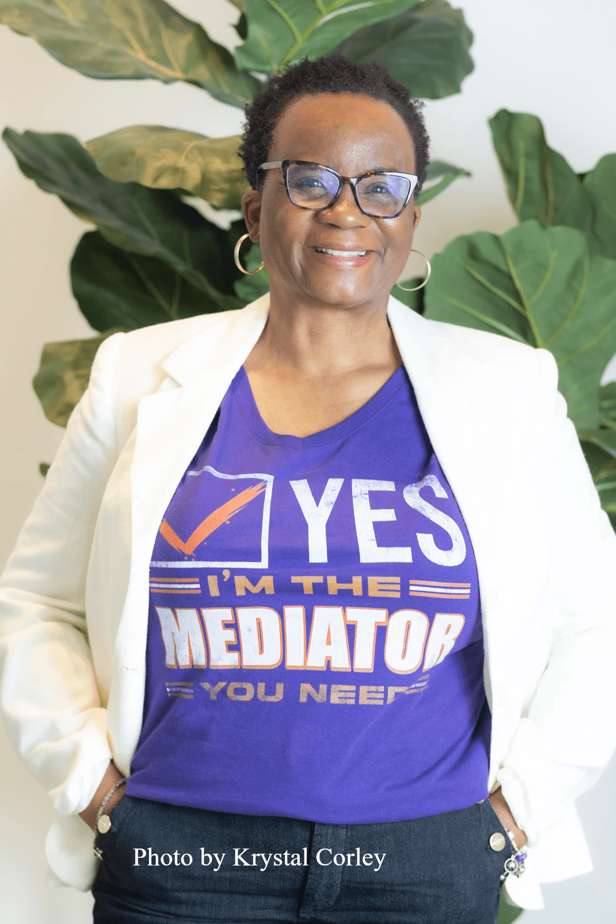 Woman with glasses smiling wearing white blazer and tshirt that says "Yes I 'm the Mediator You Need"