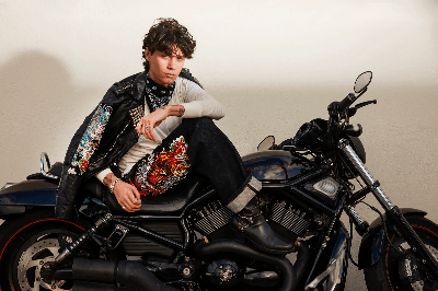 Man in a studded leather jacket and Ed Hardy tiger graphic vest sitting on a black motorcycle.
