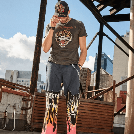 Ed Hardy men's grey tiger graphic tee with flame-print jeans and embroidered trucker hat on rooftop.