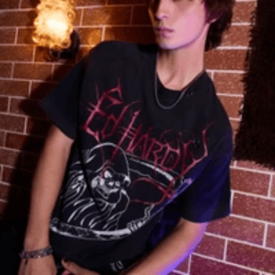 Man in a black Ed Hardy T-shirt with a red gothic skull graphic, leaning against a brick wall.