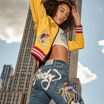 Model wearing a yellow graphic jacket and blue embroidered jeans, posing against a city skyline.