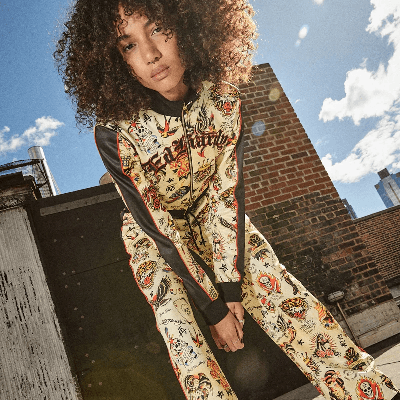 Woman on a rooftop wearing a cream and black Ed Hardy tracksuit covered in colorful tattoo-style graphics.